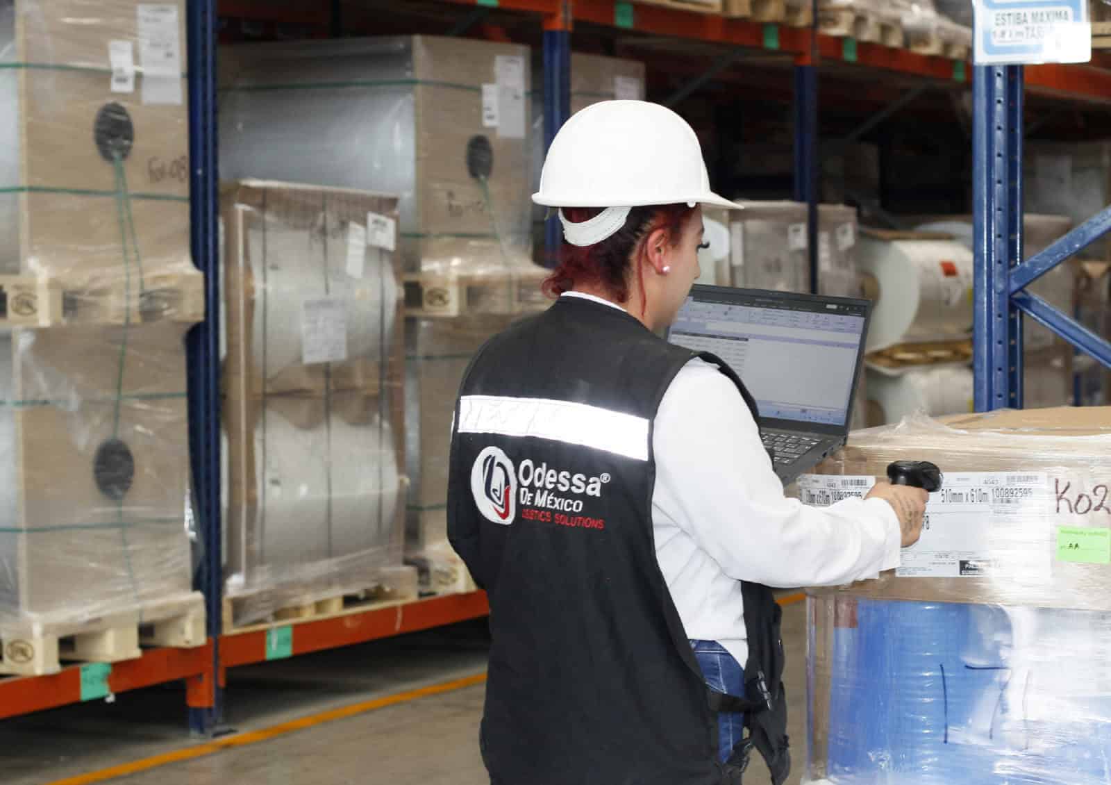 Mujer en almacén revisando inventario con laptop y equipo de seguridad.